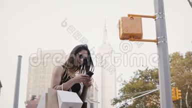 快乐美丽的白人女商人，戴着太阳镜，在帝国大厦附近使用智能手机应用程序购物